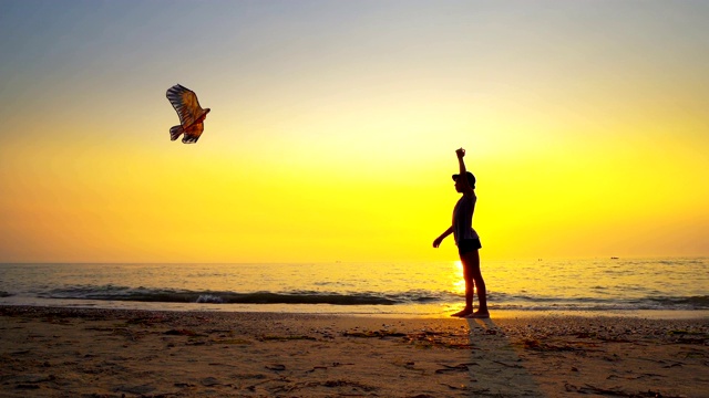 一个年轻男孩的剪影与帽子在一个空的海滩上放风筝背光的夏天日落太阳，电影镜头视频素材