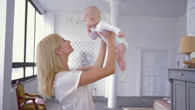 父母的爱，快乐的年轻妈妈在屋里咬着手指的可爱的小女婴的头顶上玩耍视频素材