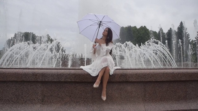 在一个阴天的喷泉边，穿着白色衣服带着雨伞的漂亮女孩视频素材