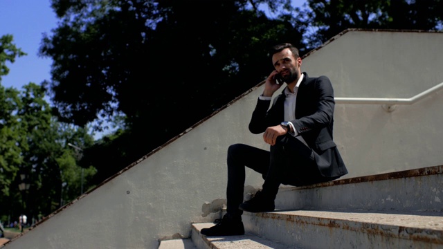 长着胡子的英俊年轻商人坐在公园的水泥楼梯上讲电话，然后看了看手上的时钟。电话洽谈业务。4 k。4 k的视频视频素材