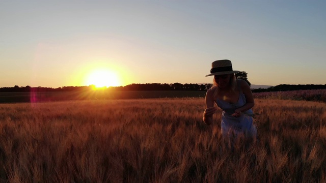 戴着帽子，穿着蓝色衣服，微笑的女游客在乡村田野上采集成熟的小麦，享受着夏日傍晚的夕阳和假期视频下载