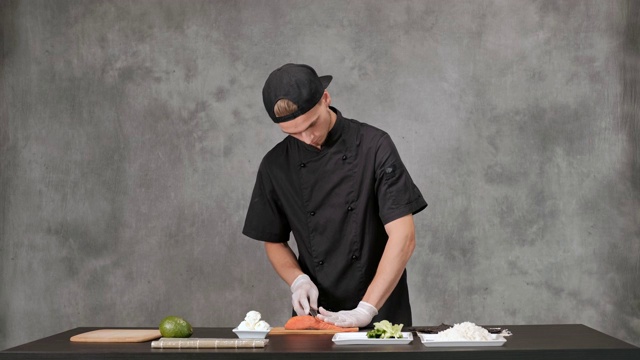 年轻的厨师厨师穿着黑色衣服在灰色的背景。厨房桌子，日本寿司和卷的原料。视频素材