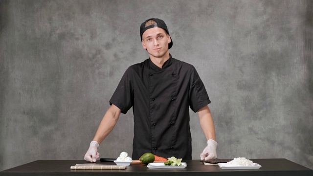 年轻的厨师厨师穿着黑色衣服在灰色的背景。厨房桌子，日本寿司和卷的原料。视频素材