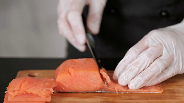 一位年轻的男性寿司厨师在桌子上用刀切着烟熏鲑鱼片。手在手套特写。视频素材