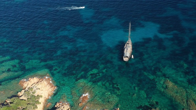 从上面俯瞰，一艘豪华帆船漂浮在美丽的绿松石海上，沐浴着撒丁岛绿色和岩石海岸。翡翠海岸(翡翠海岸)意大利。视频下载