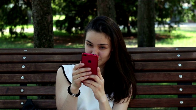 一个穿着白色t恤的漂亮小女孩正坐在公园的长椅上用智能手机在社交网络上聊天。视频素材