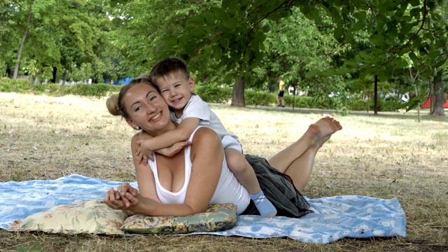 快乐的宝宝坐在妈妈的背上，而妈妈则躺在草地上，笑着拥抱着妈妈的脖子。夏天的一天，妈妈和小男孩在城市公园的草坪上玩耍。特写镜头。4 k。视频下载