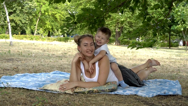 快乐的家庭:妈妈和宝宝在夏天的晚上在城市公园的草地上玩耍。小男孩坐在妈妈的背上，挑逗地笑着，拥抱妈妈的脖子，亲吻她。4 k。视频素材