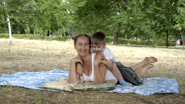 日落时分，快乐的妈妈和小儿子躺在城市公园的草坪上，微笑着对着相机摆姿势。婴儿坐在妈妈的背上，摸着妈妈的头发。他们穿着白衬衫。4 k。视频素材