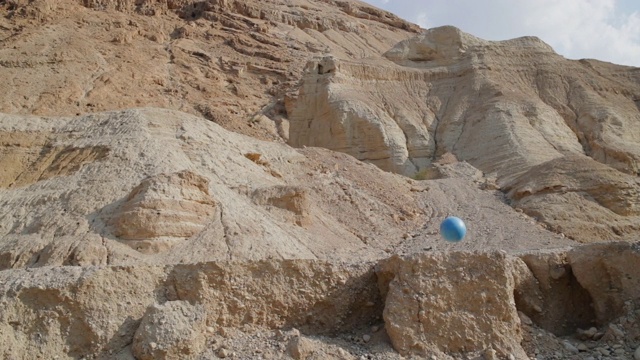慢动作:一个蓝色的运动球在一个荒凉的沙漠，以色列死海的岩石峭壁上弹跳下来视频素材