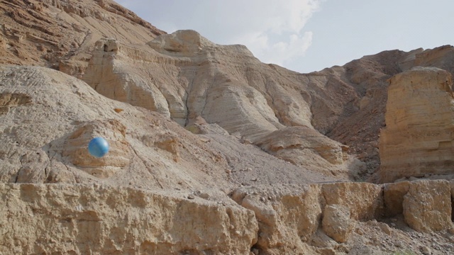 慢动作:一个蓝色的运动球戏剧性地在一个沙漠悬崖上的岩石架子上弹跳-死海，以色列视频素材