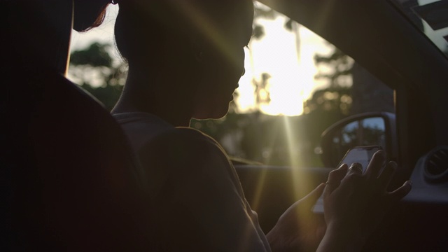 日落时分，一名长发亚洲女子在一辆汽车上用智能手机拍照视频素材