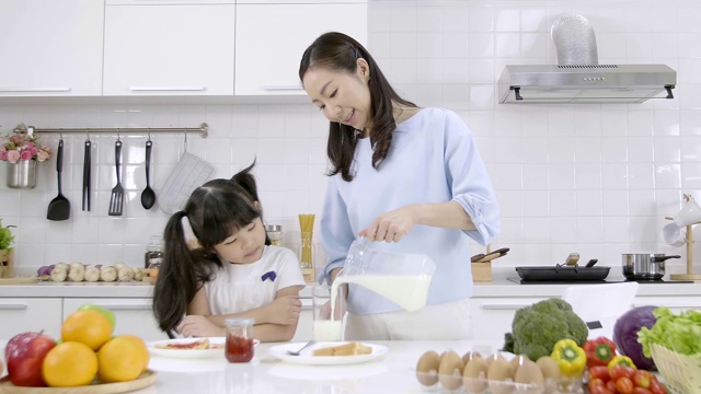 幸福的亚洲家庭在家里的厨房里做早餐。早晨妈妈把牛奶倒进玻璃杯给女儿喝。家庭烹饪的概念。慢动作视频素材