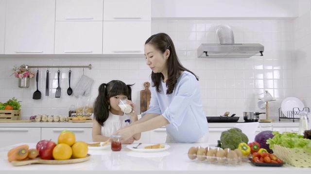 幸福的亚洲家庭在家里的厨房里做早餐。早晨妈妈把牛奶倒进玻璃杯给女儿喝。家庭烹饪概念视频素材