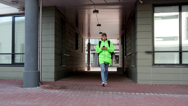 年轻的食品快递员在绿色制服和帽子走在街上的拱门与绝缘背包。用智能手机应用导航的男人视频下载