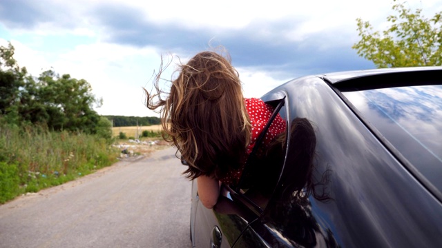 美丽的女人探出车窗，享受旅行，而骑在乡村道路。年轻的女孩看着窗外移动的汽车和她的棕色长发在风中吹拂。慢动作视频素材