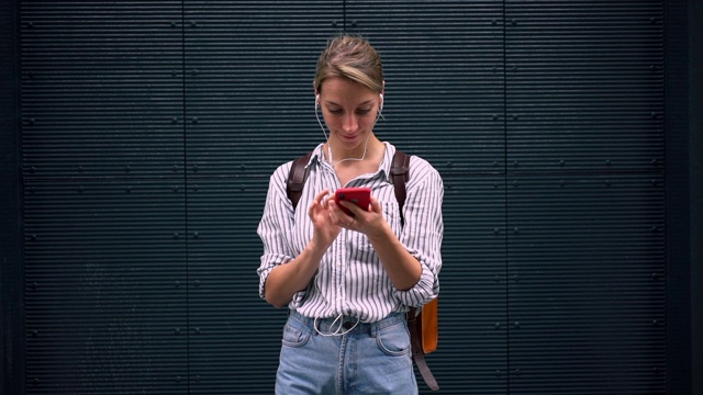 都市背景下，戴着时尚耳机的女性在智能手机上下载有声书的慢动作效果视频素材