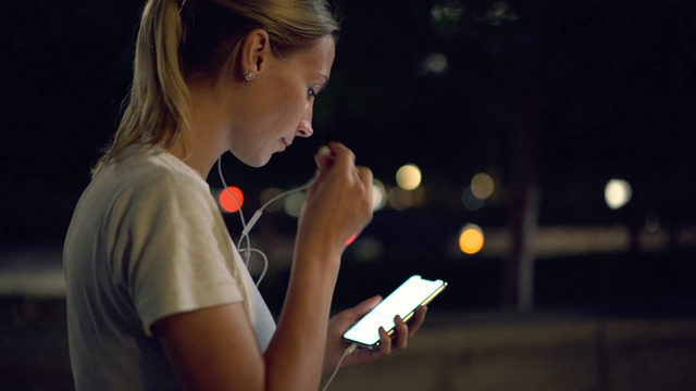 年轻一代的女孩晚上戴着电子耳机在城市街道上听音乐的慢动作效果视频素材