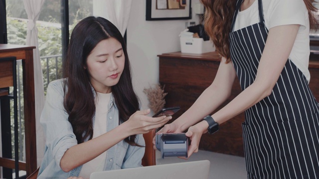 亚洲年轻女性顾客用智能手机与年轻的女咖啡师在咖啡馆的桌子上付款。咖啡师手持信用卡读卡机与顾客服务方便。视频素材