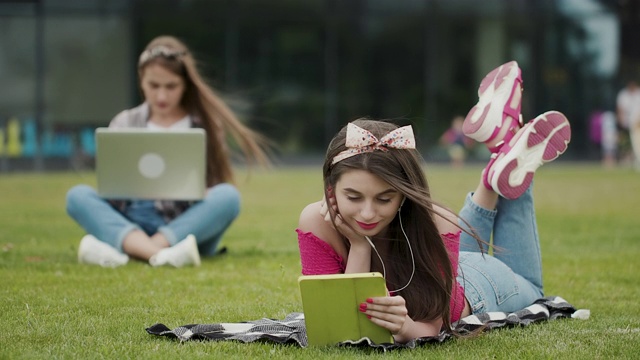 两名女性坐在一个可爱的公园的草地上，正在使用笔记本电脑和平板电脑。两个漂亮的女孩在公园里放松。视频素材