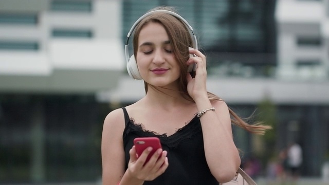 迷人的年轻女子听音乐在耳机使用智能手机在城市散步日落环顾和微笑视频素材