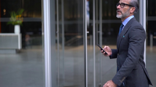 成熟的男人戴着眼镜，穿着优雅的西装，手拿着现代手机走在市中心的城市街道上视频素材