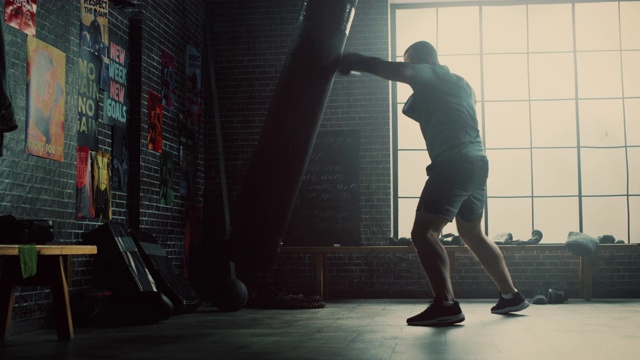 强壮和英俊的男性自由搏击手是在一个阁楼健身房与动机海报上的墙壁。男子气概的运动员正在拳击袋上练习拳击。他严肃而精力充沛。视频下载