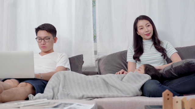幸福的亚洲家庭一起在客厅的沙发上看电视。爸爸在电脑上工作，妈妈在看电视，女儿在妈妈的腿上睡觉。视频素材
