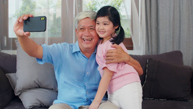 亚洲祖父和孙女在家视频通话。中国老爷爷高兴地和年轻的女孩用手机视频通话，她的爸爸和妈妈躺在客厅里。缓慢的运动。视频素材