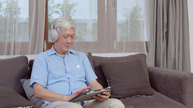 亚洲资深男士在家里放松。亚洲老年男性开心地戴着耳机，躺在家里客厅的沙发上用平板电脑听播客。视频素材