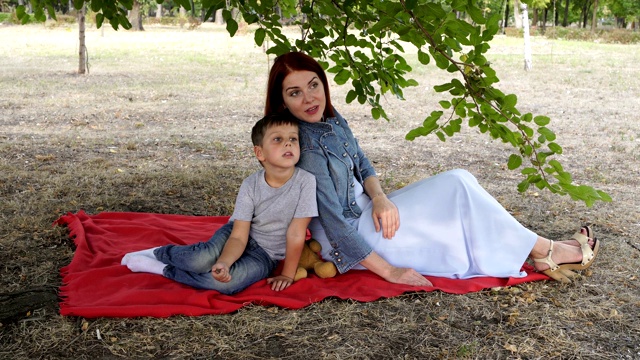 美丽的母亲和孩子在城市公园里摆姿势，肩并肩坐在绿色树枝下的红毯上。他们移开目光，微笑着。特写镜头。4 k。视频素材