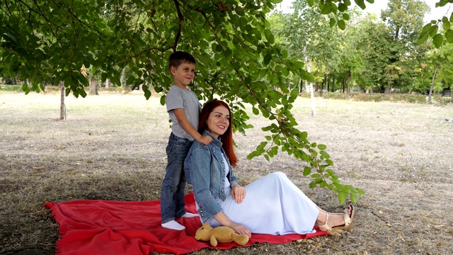 一个可爱的孩子和他的母亲坐在城市公园的绿色树枝下的红毯上。他们微笑着，在秋天的夜晚玩耍。4 k。视频素材