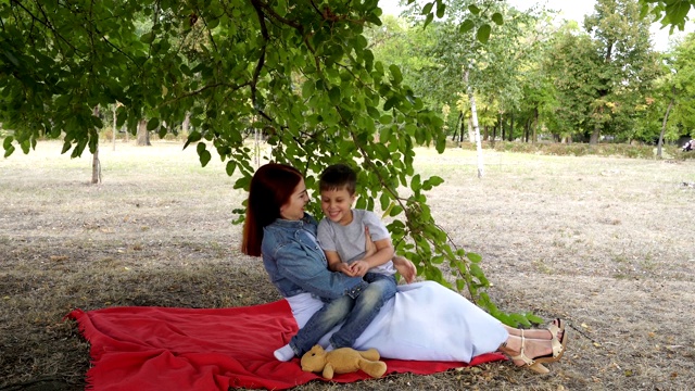 妈妈和小男孩坐在城市公园的草坪上玩耍，坐在绿色树枝下的红毯上。他们微笑着，在秋天的夜晚玩得很开心。4 k。视频素材
