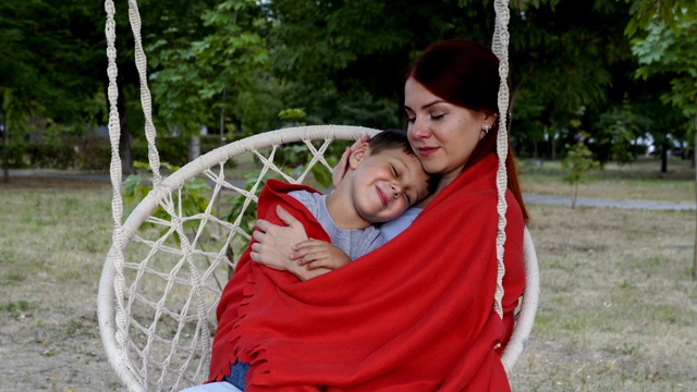 一位快乐的母亲在日落时分坐在公园的秋千上抱着她的小儿子。她把它裹在红毯子下面，抚摸着它。的概念。特写镜头。4 k。视频素材