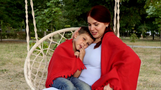 一个快乐的妈妈和婴儿在日落公园的秋千上的肖像。一个红头发的女孩把年幼的儿子抱在膝盖上，拥抱他，给他盖上红毯子。关闭了。4 k。视频素材