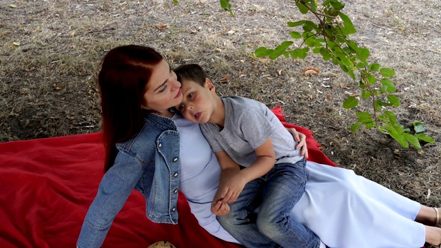妈妈抱着她的小儿子坐在城市公园的草坪上，坐在绿色树枝下的红毯子上。孩子躺在妈妈的肩膀上，妈妈抚摸着他，微笑着。概念。4 k。视频素材