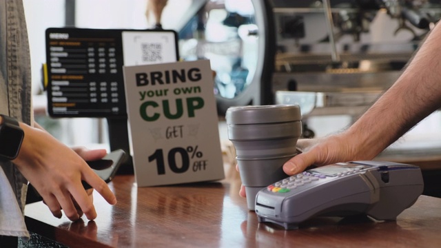 零浪费的生活理念。亚洲女性顾客带着自己的杯子买咖啡，用手机非接触式支付给柜台吧台的咖啡师的慢动作视频下载