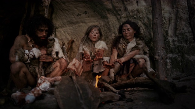 穿着兽皮的史前狩猎采集者部落晚上住在洞穴里。尼安德特人或智人家庭试图在篝火取暖，手在火上，烹饪食物视频下载