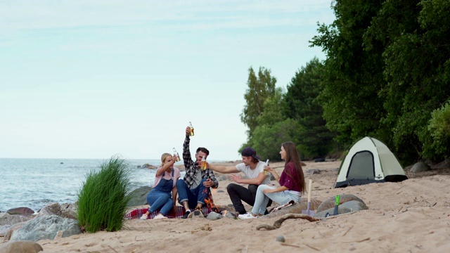 广角拍摄的一群快乐的年轻朋友，两男两女，在海滩上的篝火上用啤酒瓶敬酒，他们的帐篷在背景视频素材