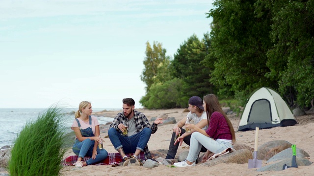 四个年轻人在海滩上露营。夏日里，两男两女围坐在营火旁，喝着啤酒聊天视频素材