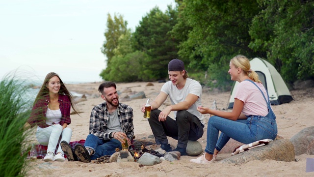 四个年轻人在海滩上露营。两男两女坐在营火旁，喝着啤酒有说有笑视频素材