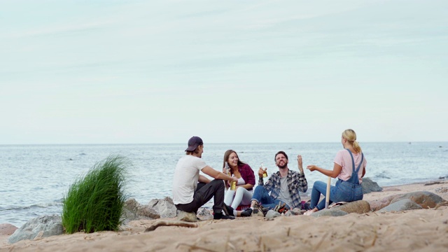一组四个年轻的朋友围坐在海边的海滩篝火玩。两个男人和两个女人一边喝着啤酒，一边有说有笑视频素材