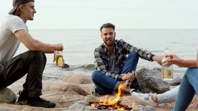四名年轻人在海边的海滩上围坐在篝火旁放松。快乐的年轻人和朋友聊天视频素材