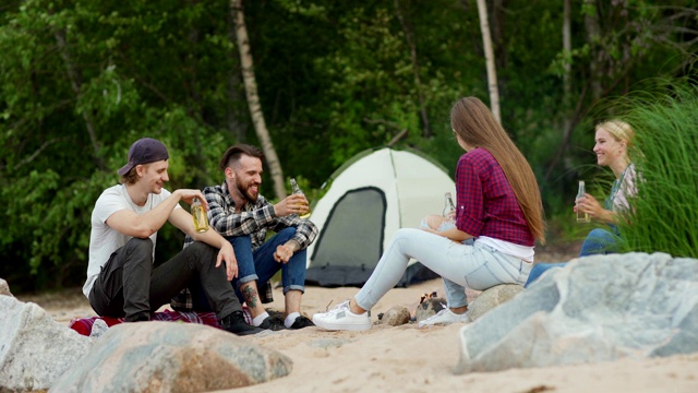 四个快乐的年轻朋友在享受露营。两个男人和两个女人在篝火旁喝啤酒聊天视频素材