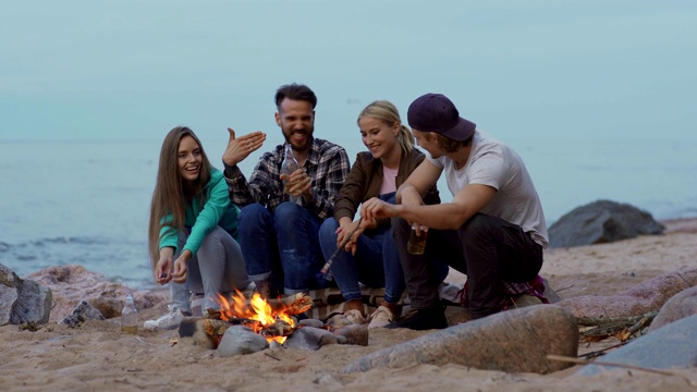 四个年轻人拿着啤酒瓶在海滩上的篝火旁放松。一个愉快的年轻人在给他的朋友讲有趣的故事视频素材