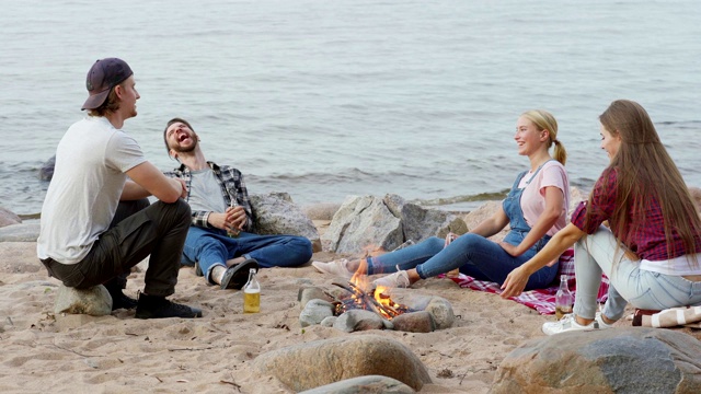四个年轻的朋友，两男两女，围坐在海滩上的篝火旁，坐在岩石上，喝着啤酒，愉快地聊天。年轻人在笑话后大笑视频素材