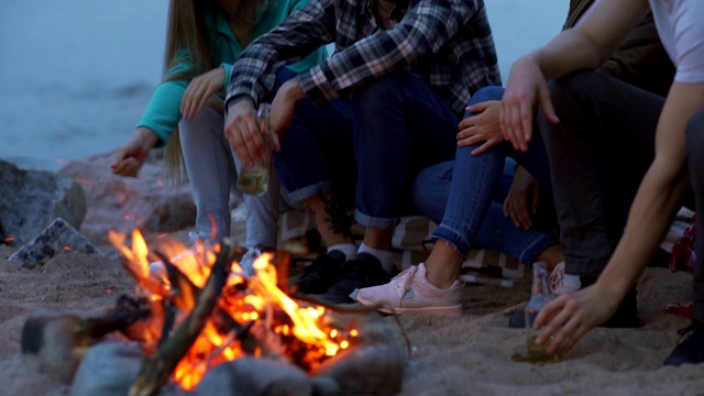 倾斜镜头，四个年轻的朋友，两男两女，坐在海滩上的篝火旁，喝啤酒，在晚上有说有笑视频素材