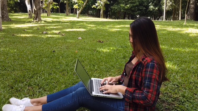 商务亚洲女性使用笔记本电脑工作在笔记本电脑键盘上打字和坐在公园的绿色草地在户外。是对焦不够或对焦太软。视频素材