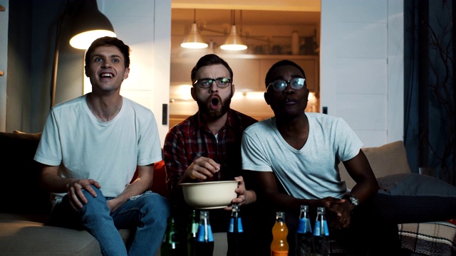 快乐的多民族青年男子观看体育比赛，一起在家里用大放映机屏幕上的零食加油慢动作视频素材