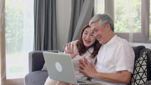 亚洲老年夫妇在家视频通话。中国爷爷奶奶，躺在家里客厅的沙发上，用笔记本电脑视频通话与家里的孙子孙女聊天。缓慢的运动。视频素材